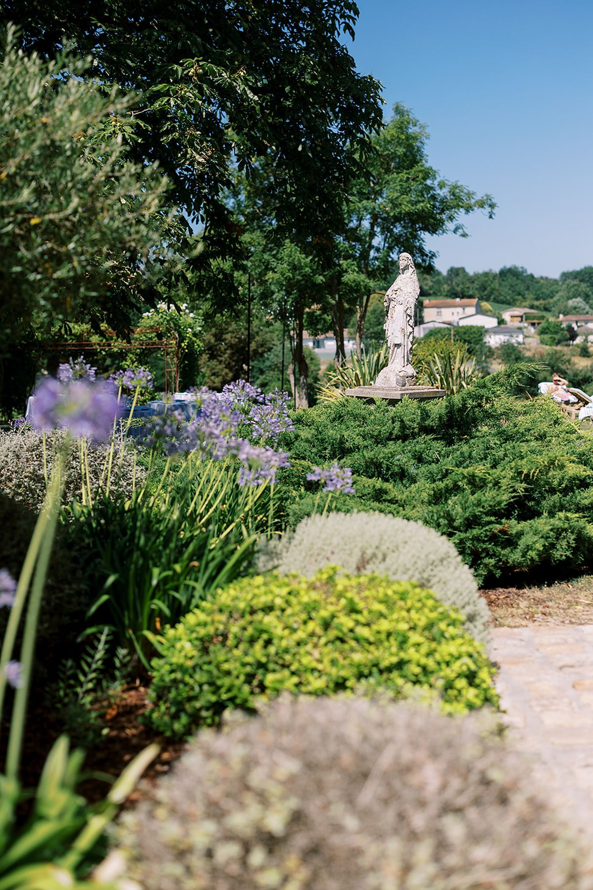 A sunlit garden with rounded clipped shrubs, blue agapanthus flowers and a stone statue, with countryside beyond.