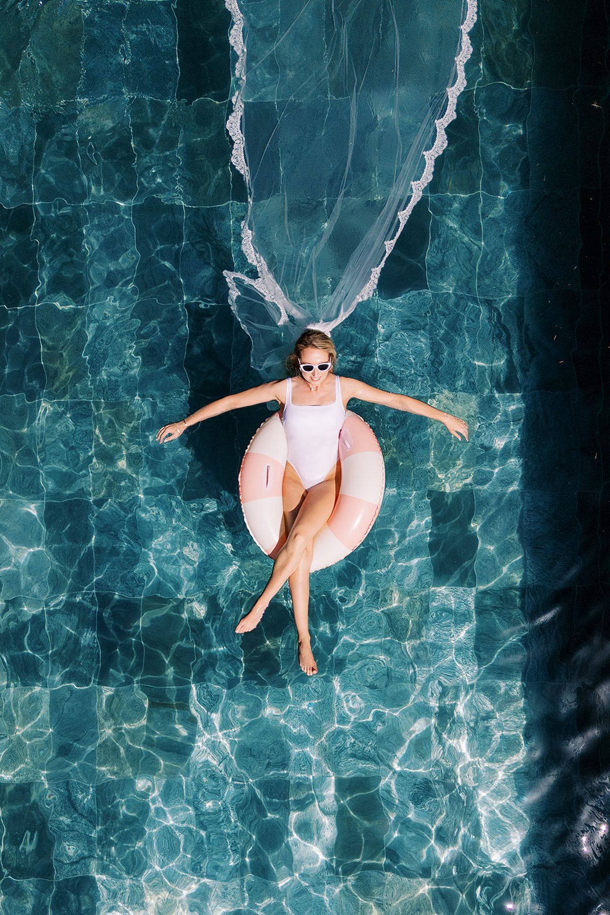 Seen from above, a woman in a white swimsuit and sunglasses floats on a pink ring in a pool, her veil trailing.