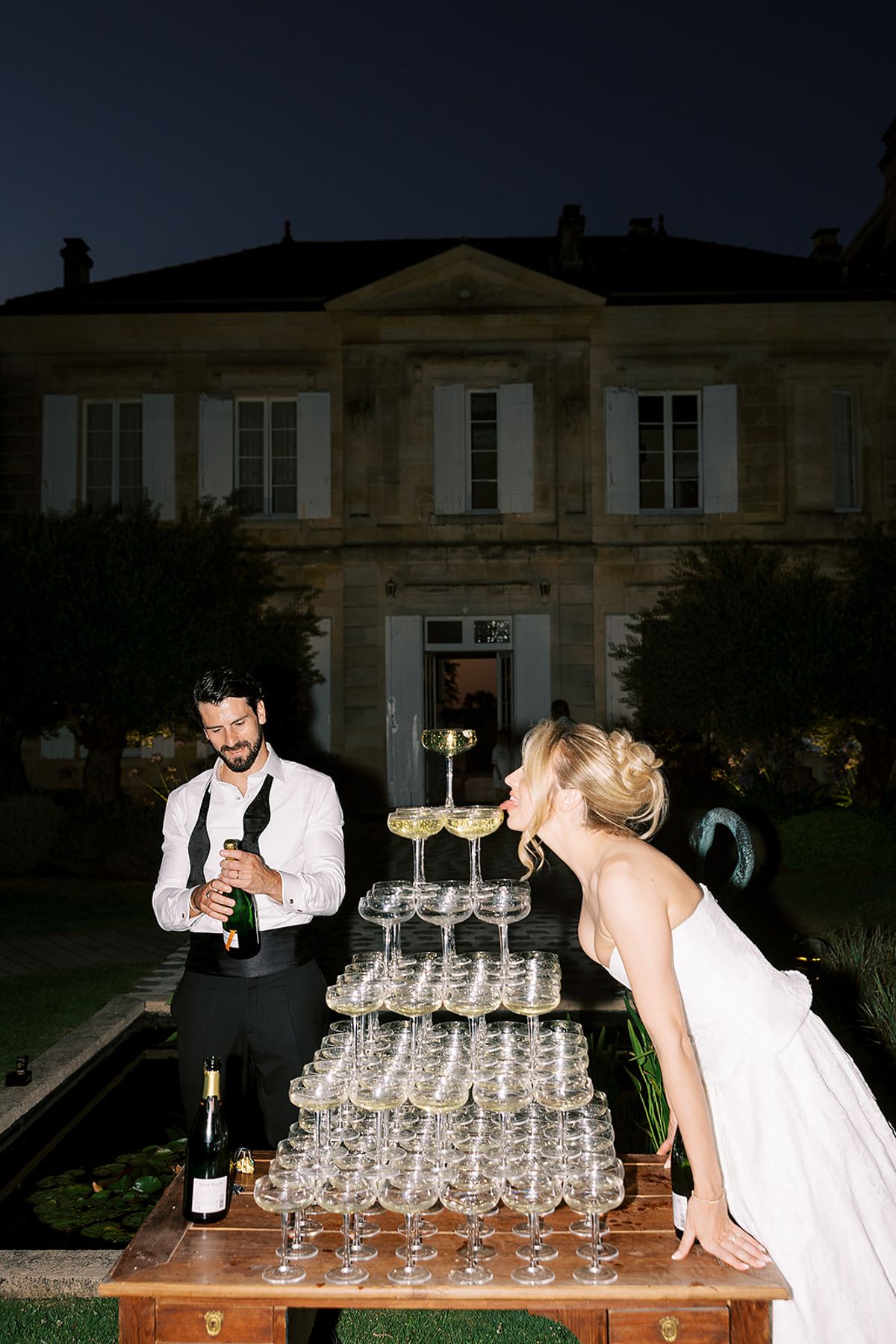 At night the groom holds a bottle beside a coupe tower while the bride leans in towards the glasses, the chateau lit behind.