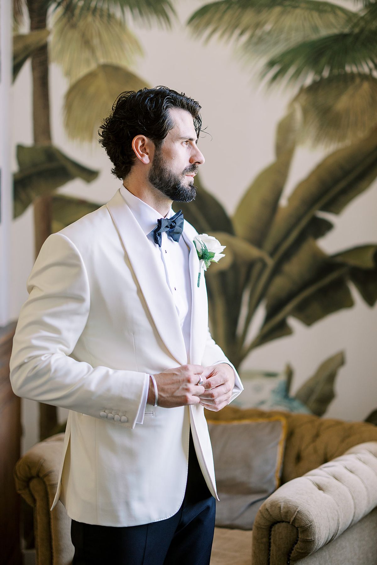 A bearded man in a white dinner jacket and bow tie looks to one side, standing in front of palm leaf wallpaper.