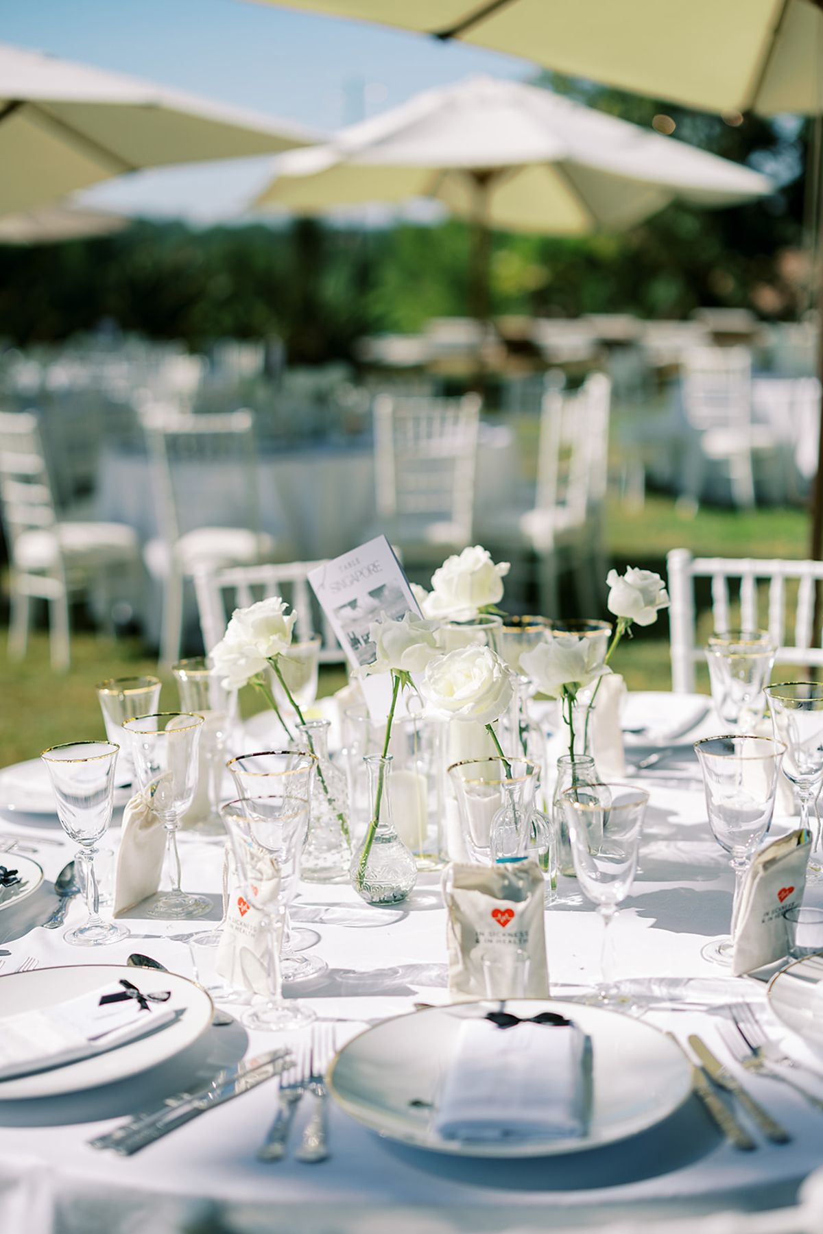 Round tables laid with white linen, glassware and single flower stems, surrounded by white chairs under cream parasols outdoors.