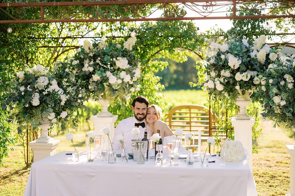 The couple sit together at a long white-clothed table beneath a pergola draped with white flowers, with vines stretching behind them.