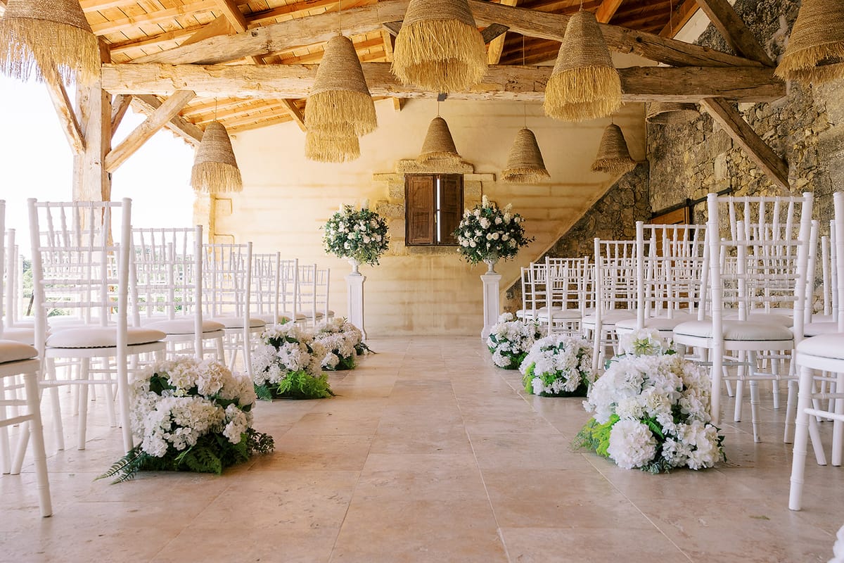 Empty ceremony aisle in a rustic stone barn, lined with white chairs, white flower arrangements and hanging straw lampshades.