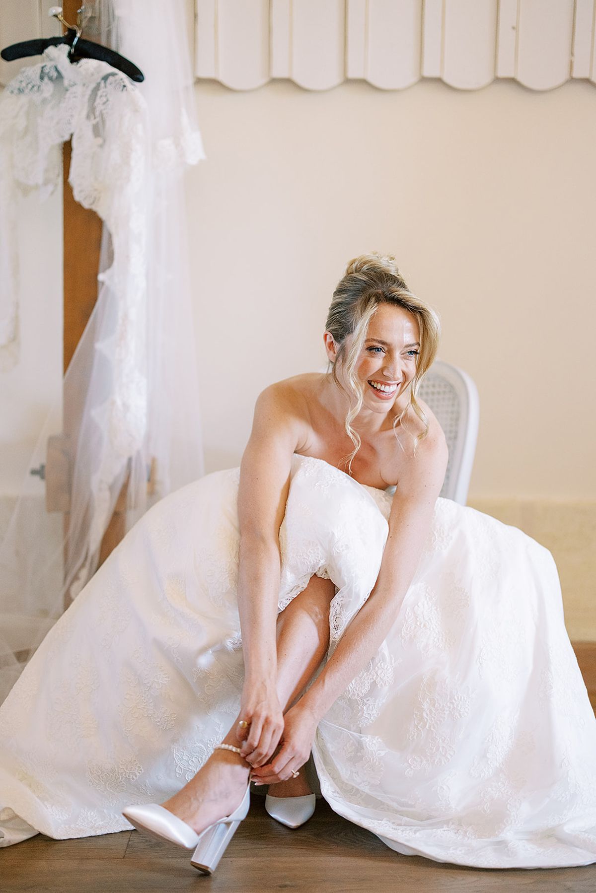 The bride sits in her full-skirted gown, smiling as she fastens a white shoe, with a dress hanging behind her.