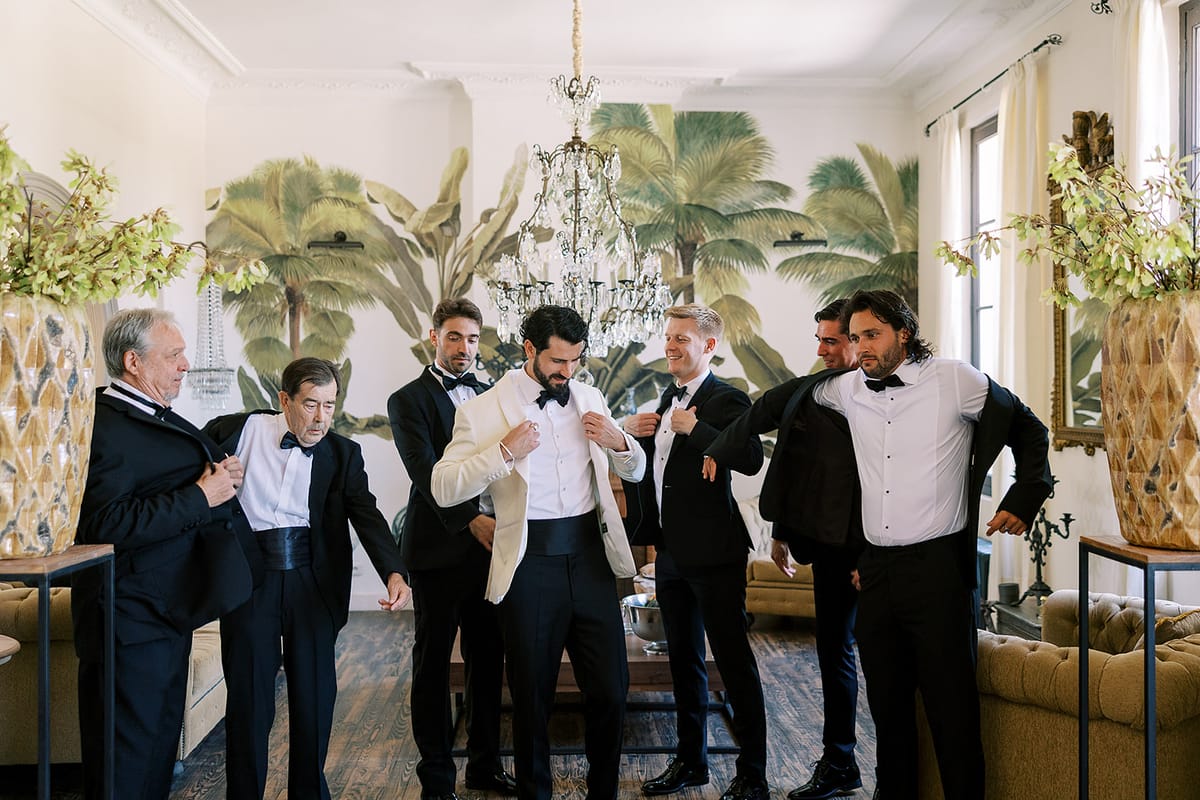 Six men in black tie and one in a white jacket help each other dress in a bright room with palm wallpaper.
