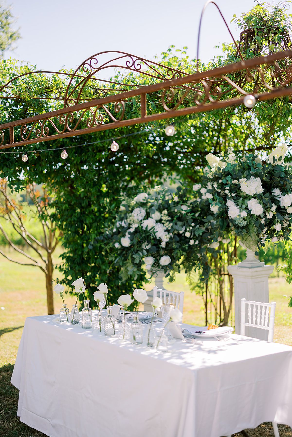 A white-clothed table with small vases sits beneath a rusty metal arch hung with festoon lights and white blooms.