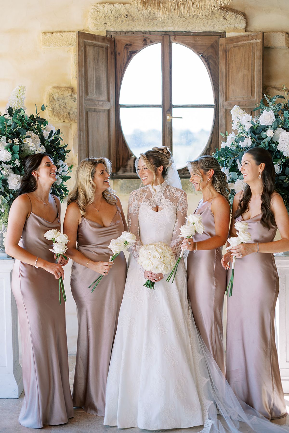 The bride in a lace-sleeved gown stands among four bridesmaids in blush satin dresses, all holding bouquets and smiling.