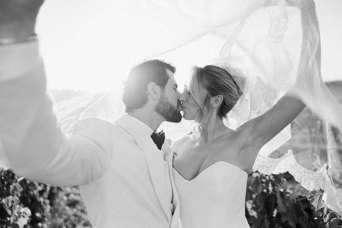 In black and white, the groom and bride kiss as a veil lifts in the wind around them.