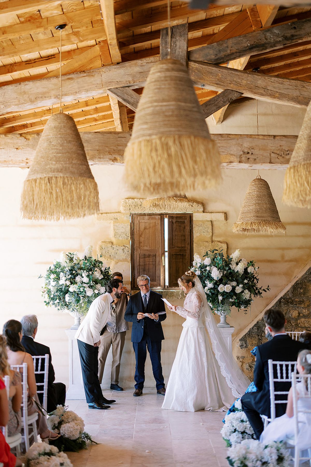 The couple stand before an officiant beside white flower arrangements while seated guests watch, beneath beams and woven pendant lights.
