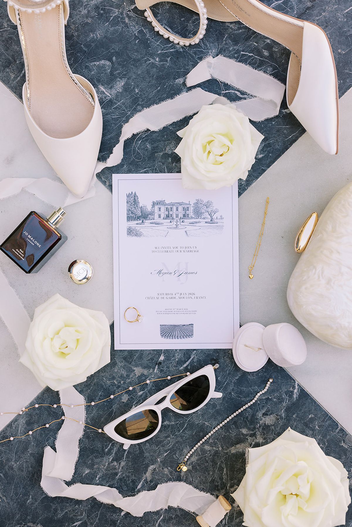 Seen from above, an illustrated invitation, white heels, white roses, sunglasses, perfume and ribbon lie on dark marble.