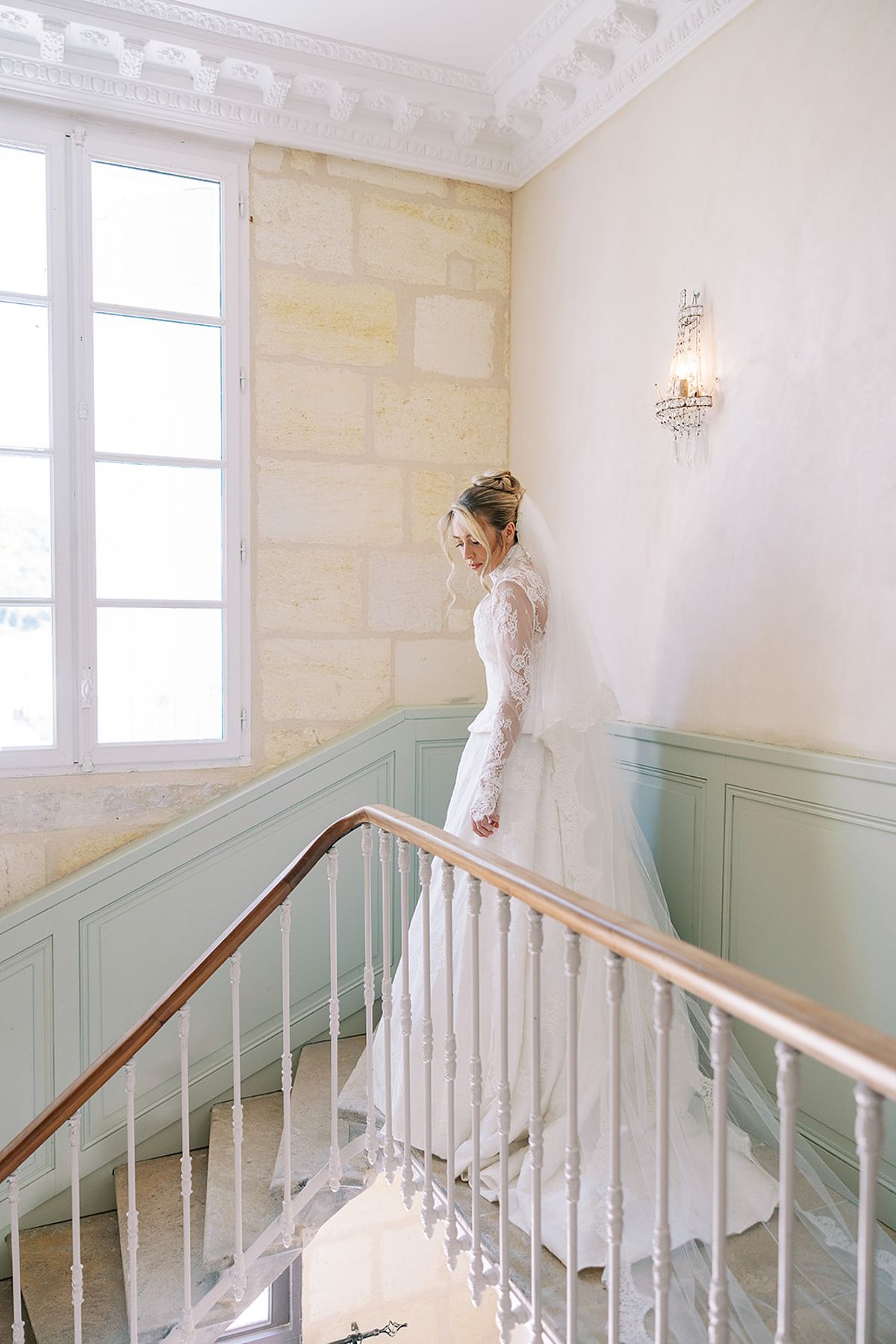 The bride, seen from behind in a long-sleeved lace gown and veil, stands on a staircase with a metal banister beside a tall window.