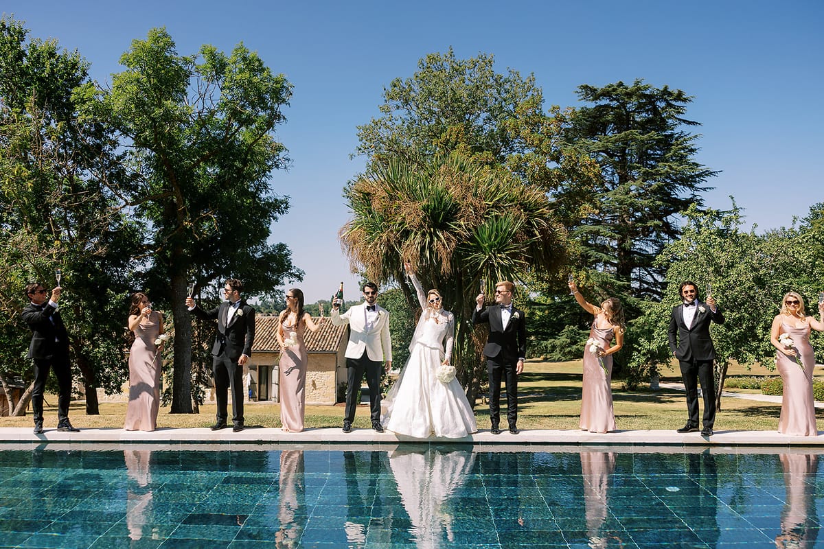 The couple stand centrally with bridesmaids and groomsmen on either side along the far edge of a pool beneath tall trees.