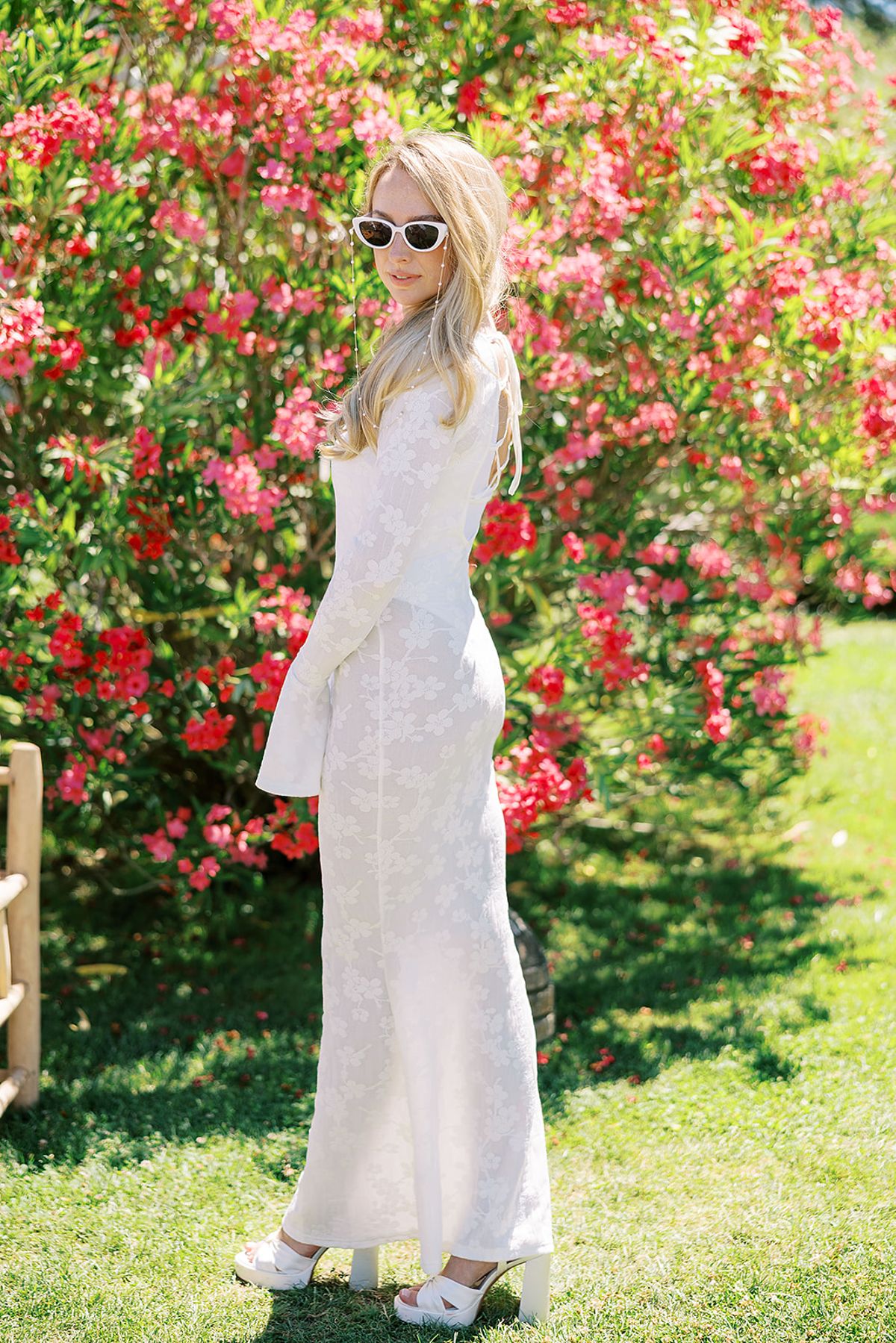 The bride in a long white outfit and white sunglasses stands on the grass in front of a bush of bright pink flowers.