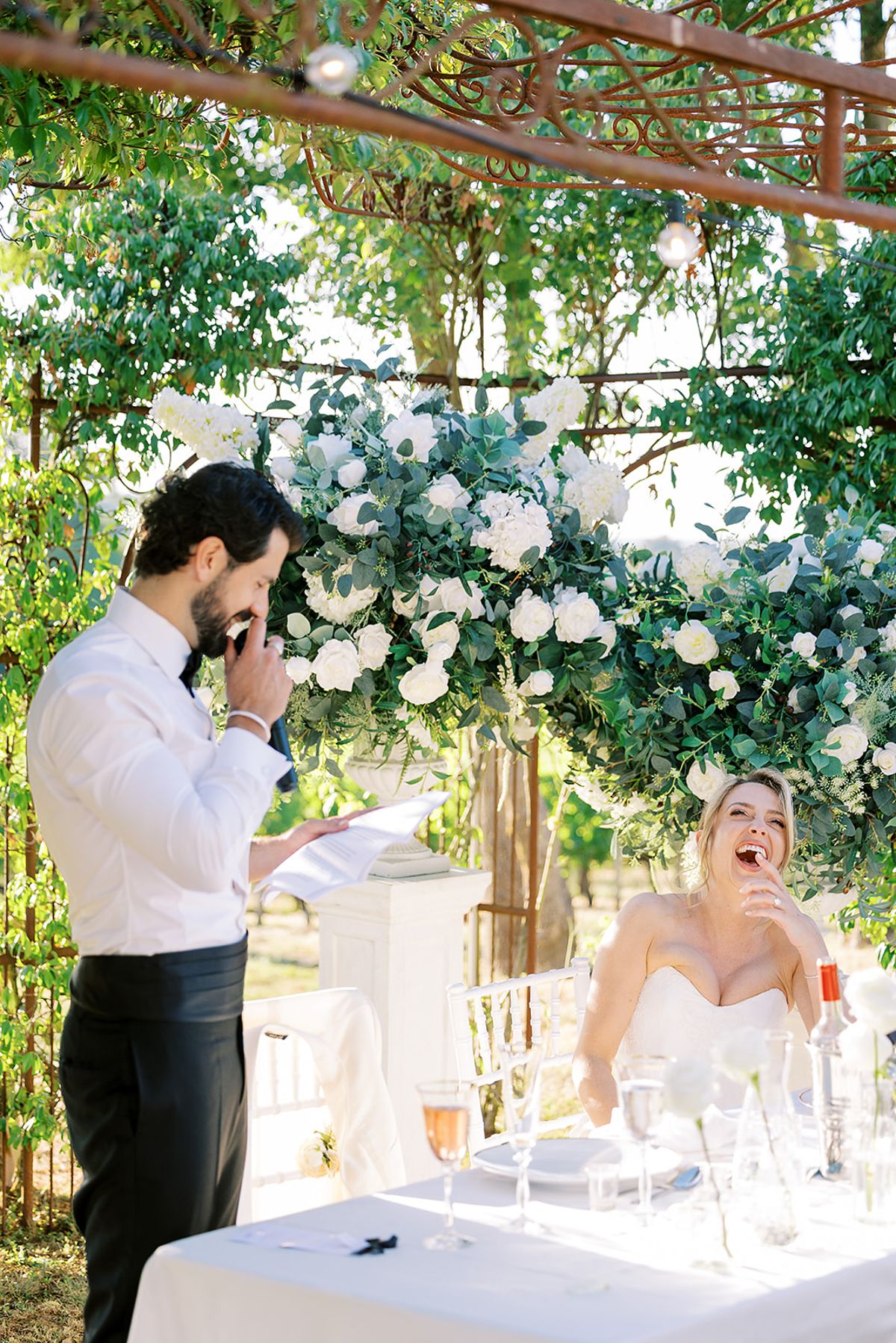 The groom reads from a sheet at a lectern, hand to his face, while the bride laughs at a table beneath white flowers.
