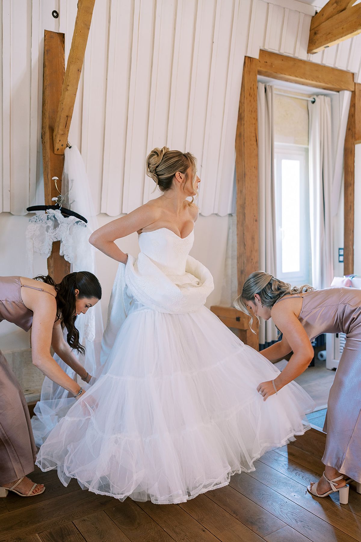 Two bridesmaids crouch to arrange the bride's tulle skirt as she stands in a bright room with wooden beams.