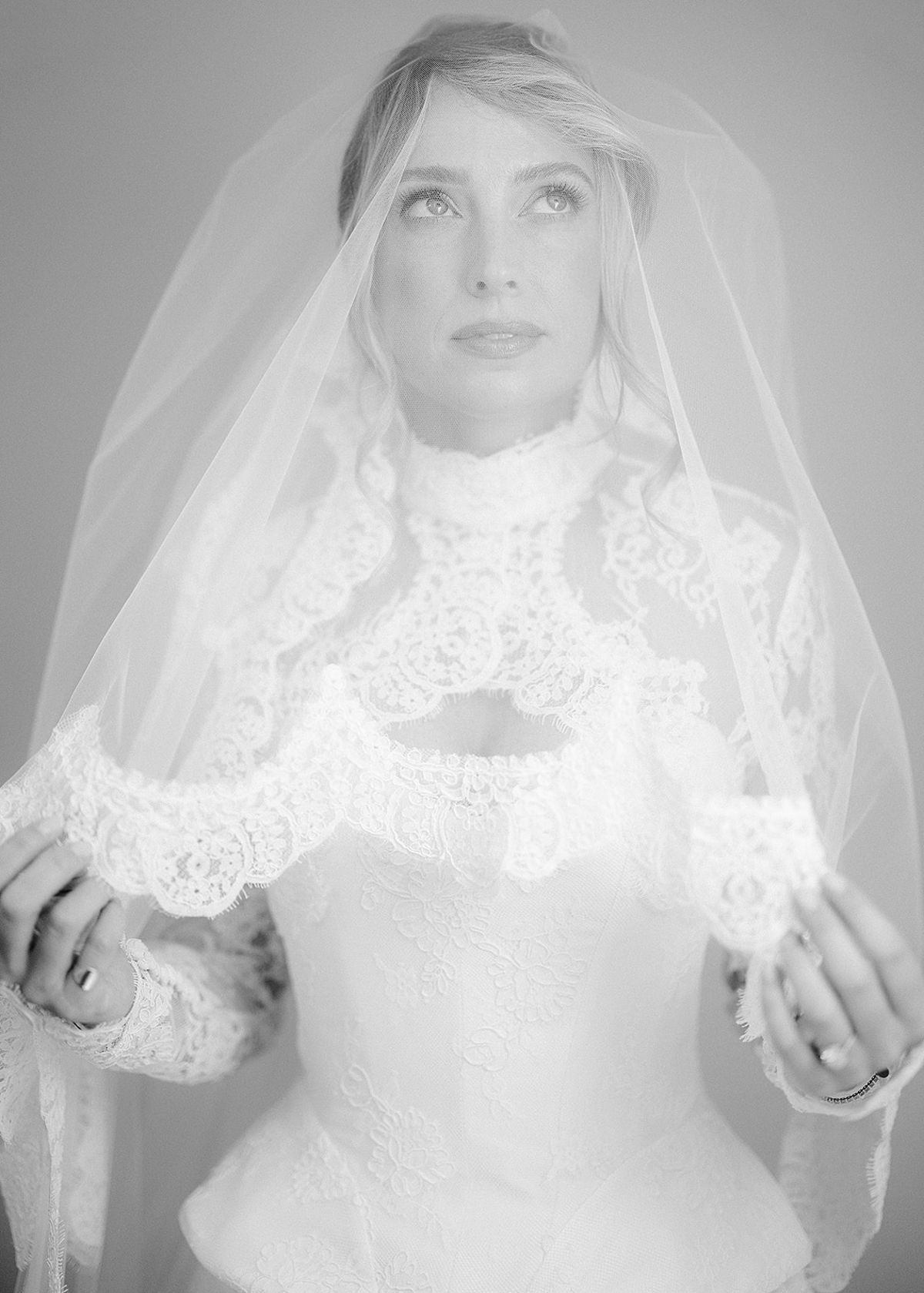 The bride gazes upwards, holding a lace-edged veil in front of her high-necked lace gown, in black and white.