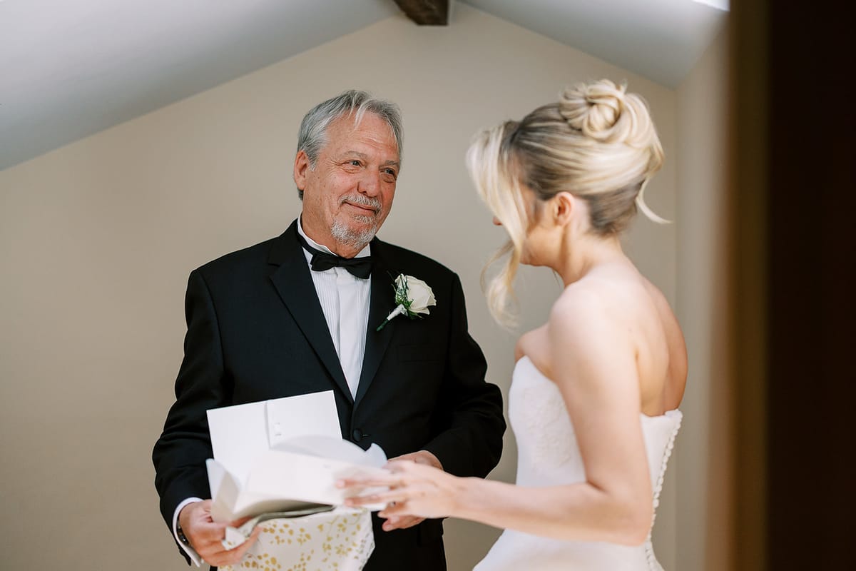 An older man in a black tuxedo holds a card and an envelope while looking at the bride, whose hair is in an updo.