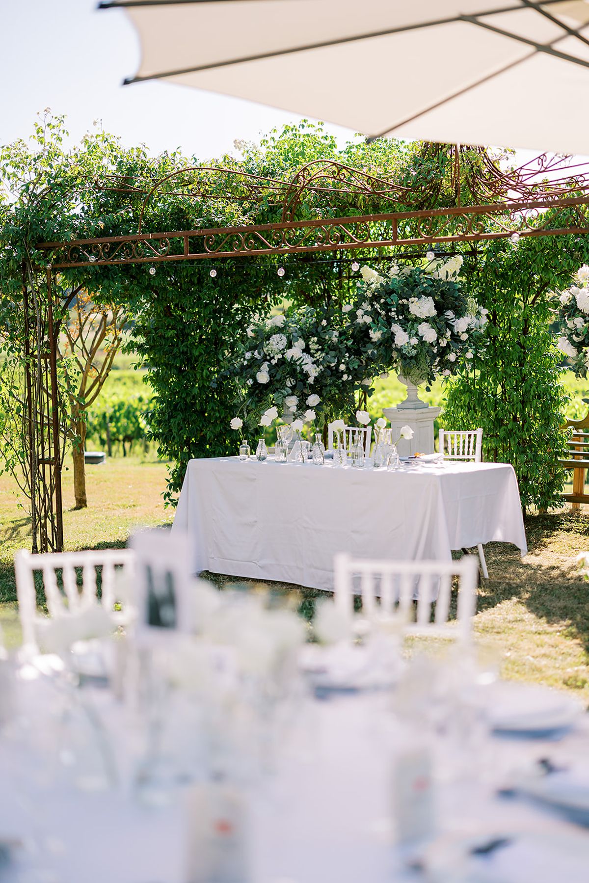 A table with a white cloth is set beneath a pergola covered in greenery and white flowers, with blurred glassware in the foreground.