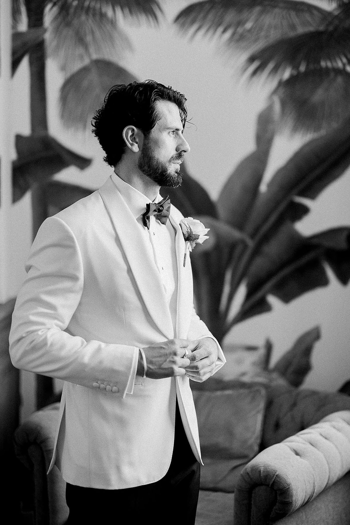 In black and white, the groom in a white dinner jacket and bow tie looks aside while adjusting his cuff, with leafy wallpaper behind.