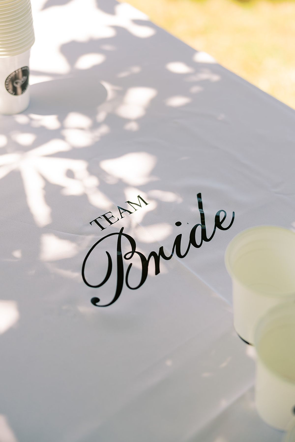 The words Team Bride in black script lettering appear on a pale surface speckled with shadows, beside a small cup.