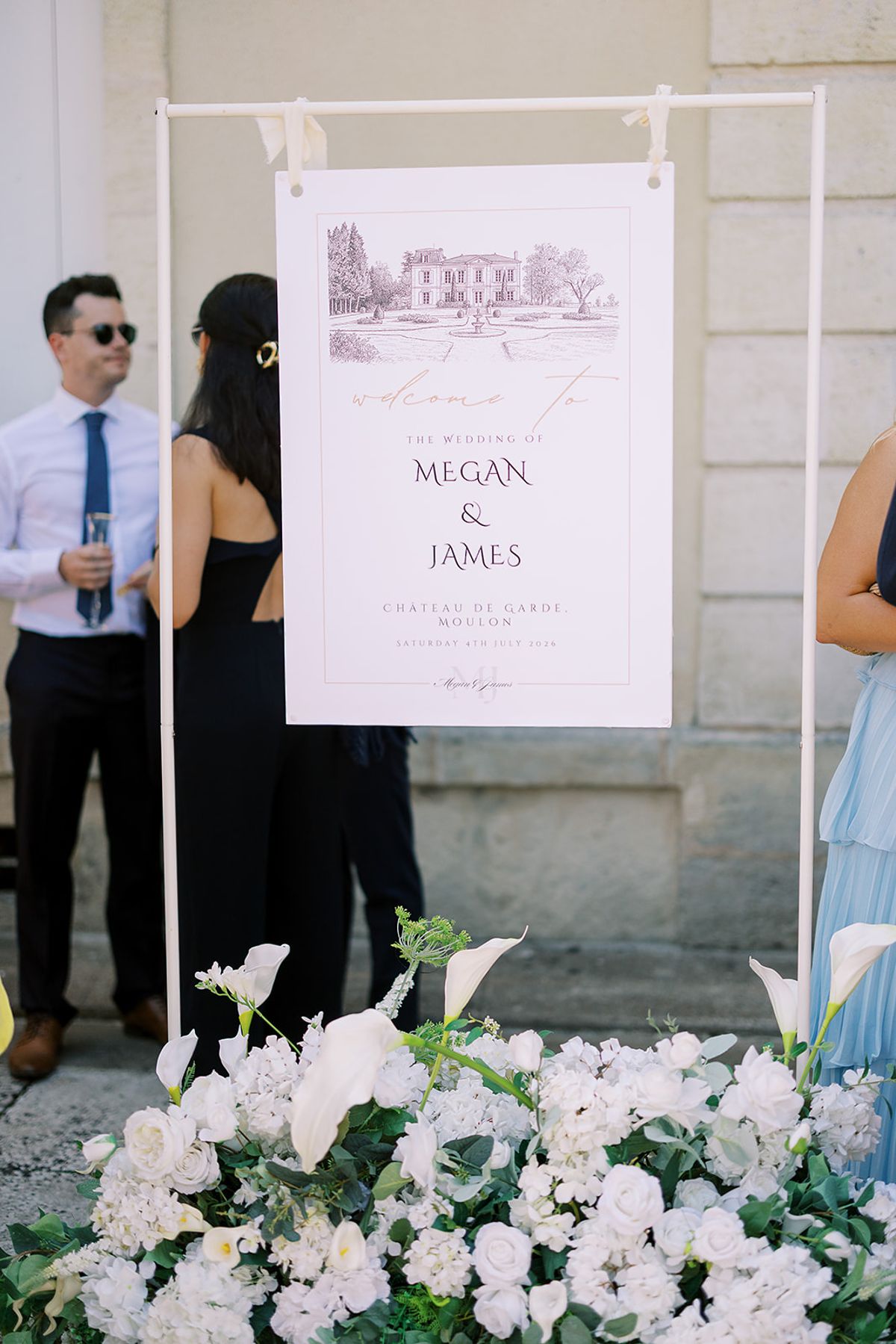 A framed illustrated welcome sign stands above an arrangement of white flowers, with guests chatting beside it.