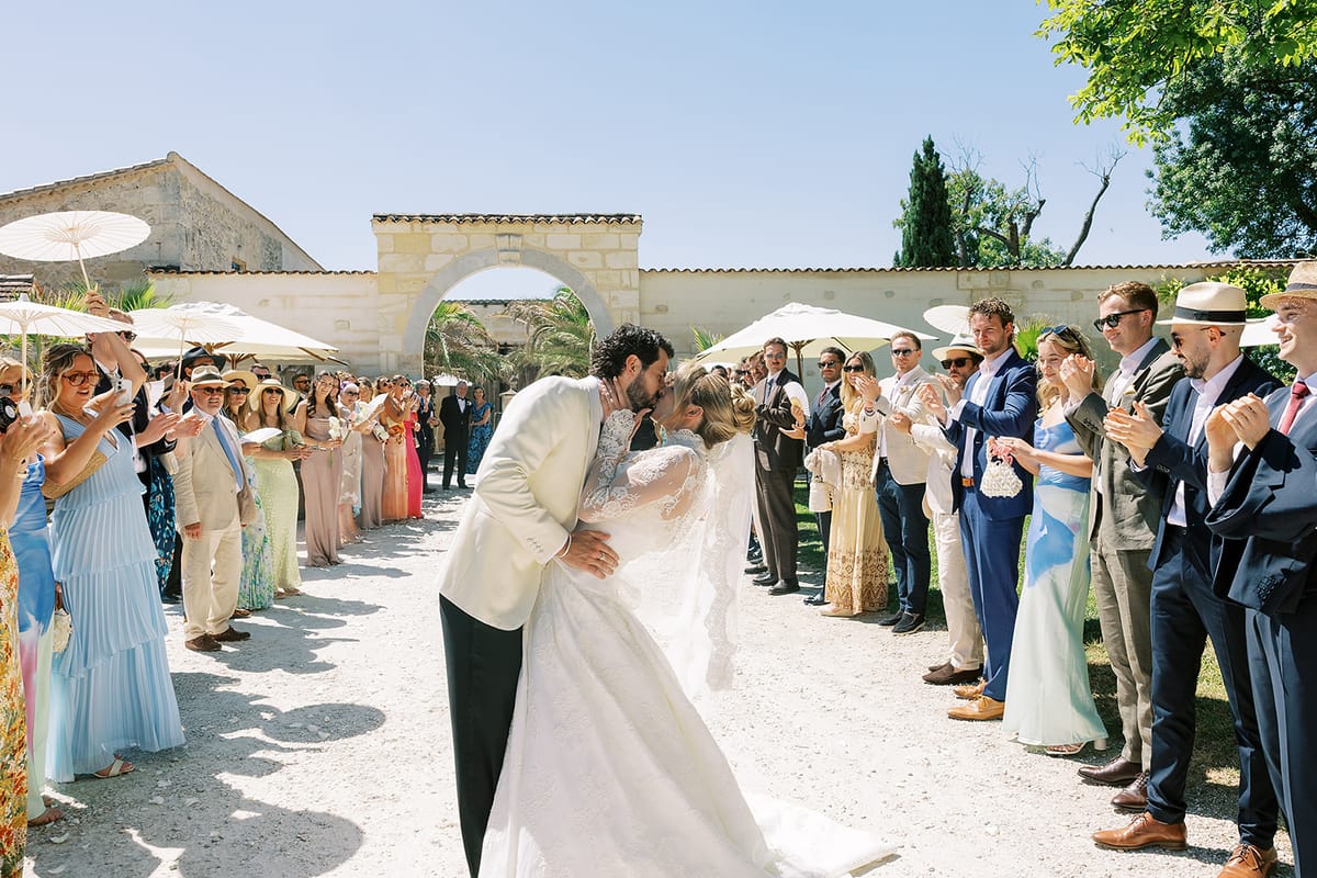 The couple share a kiss on a gravel path lined on both sides by guests in colourful outfits, in a stone courtyard under a blue sky.