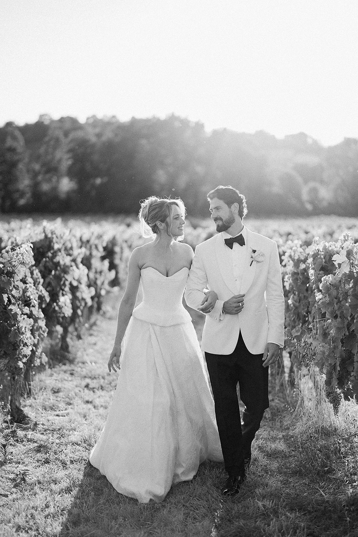 In black and white, the couple walk arm in arm between vine rows, smiling at each other in soft backlight.