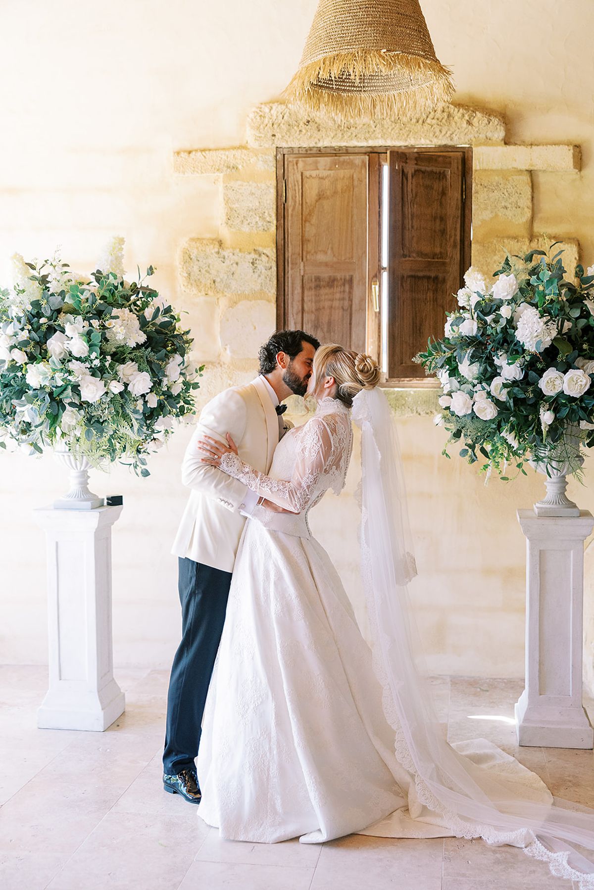 The couple kiss in front of a wooden shuttered window, between two white flower arrangements, with the bride's veil trailing behind.