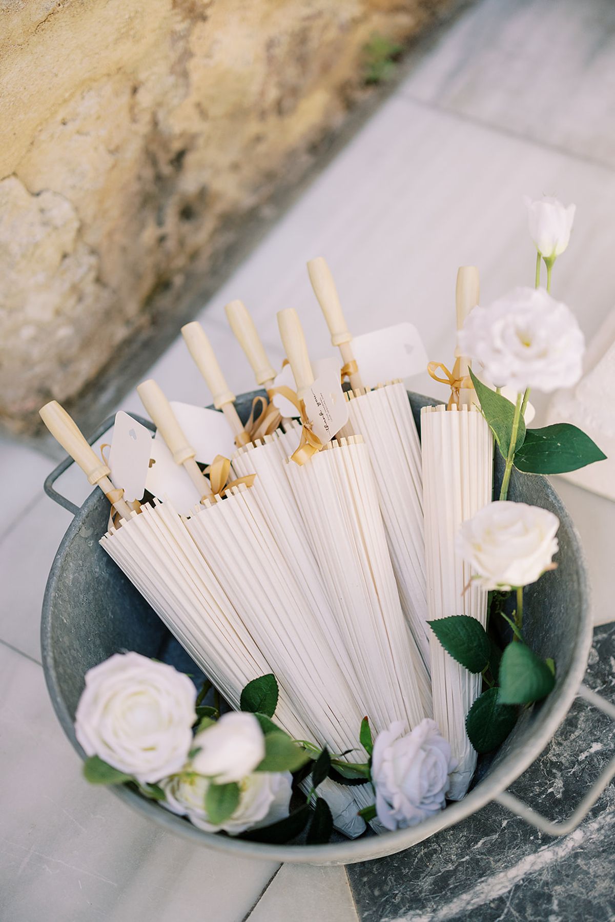 A galvanised bucket holds cream paper parasols with wooden handles and white roses, seen from above on a stone floor.