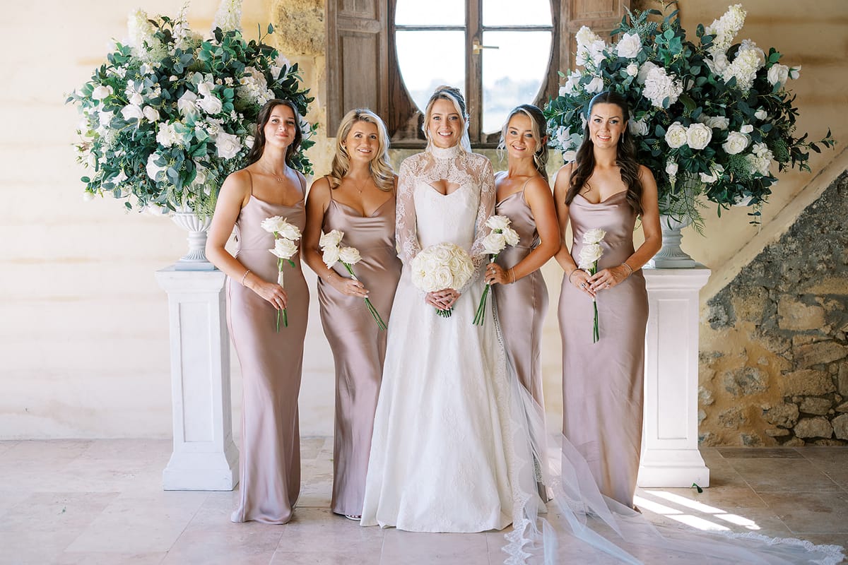 The bride stands between four bridesmaids in blush satin dresses, holding white bouquets, with large floral arrangements either side.