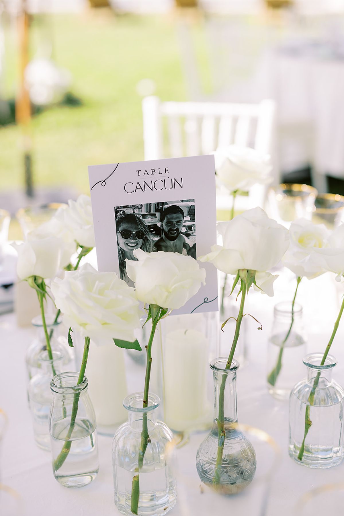 A printed table number card stands among white roses in slim glass bottles, with a pillar candle on a white tablecloth.