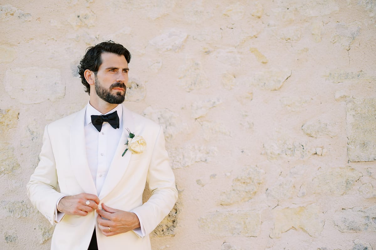 The groom, bearded and wearing a white dinner jacket and bow tie, adjusts his jacket against a pale stone wall.
