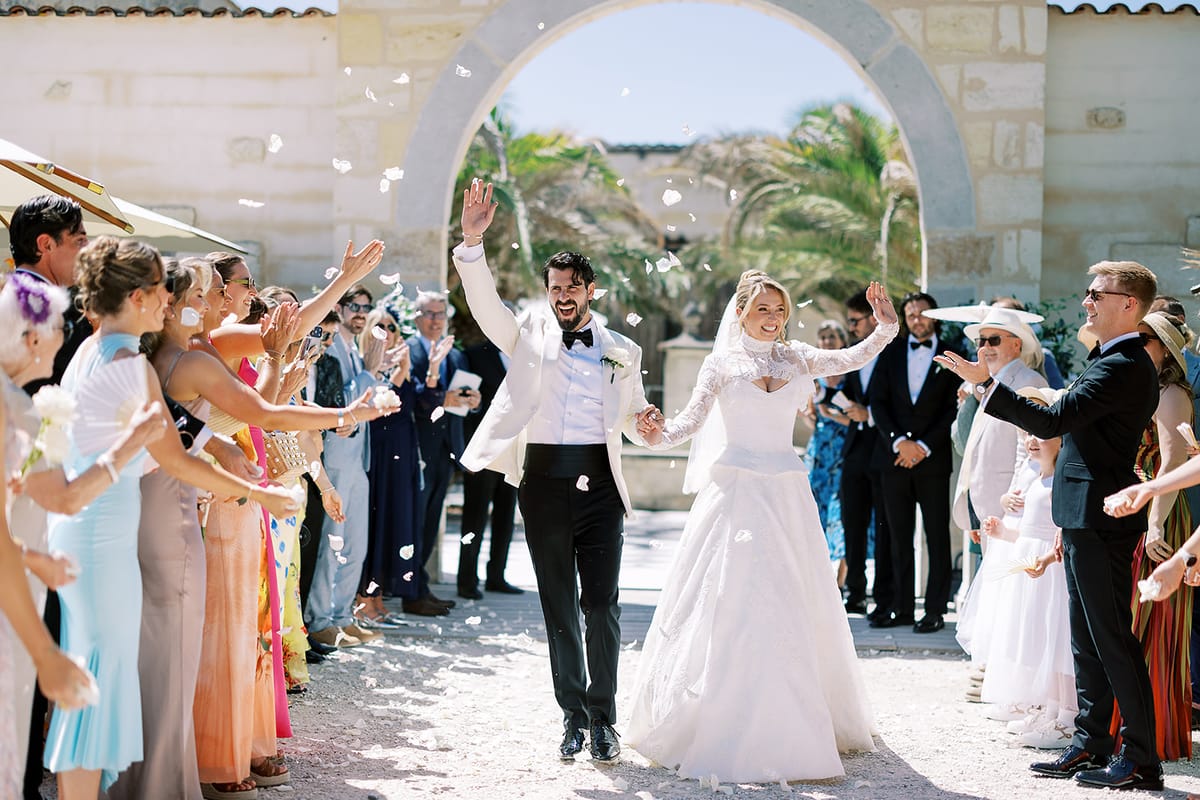 The couple walk arm in arm beneath a stone arch as guests on both sides cheer and throw confetti.