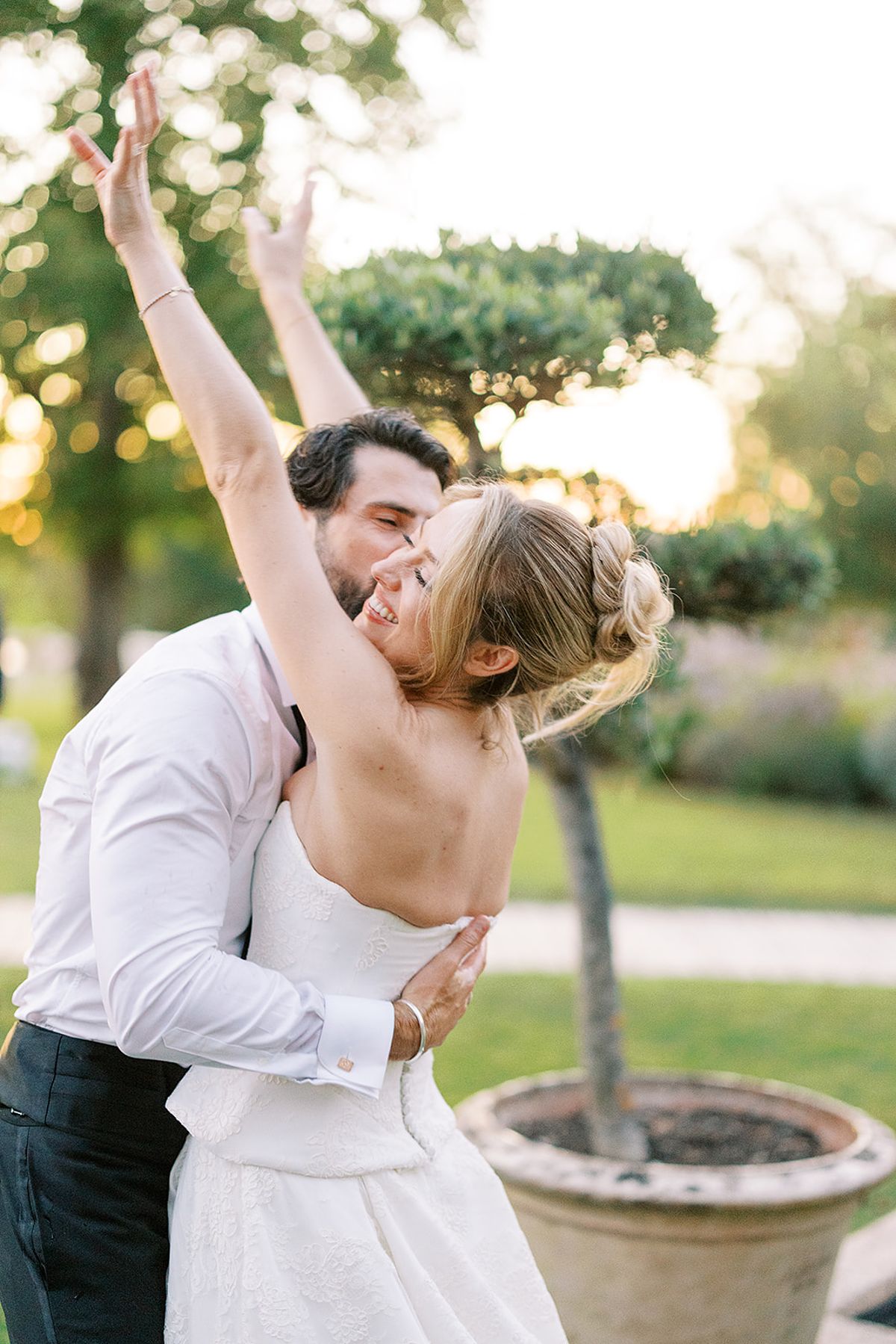 The groom in a white shirt holds the bride close as she throws her arms in the air, in a sunlit garden with a clipped tree behind.