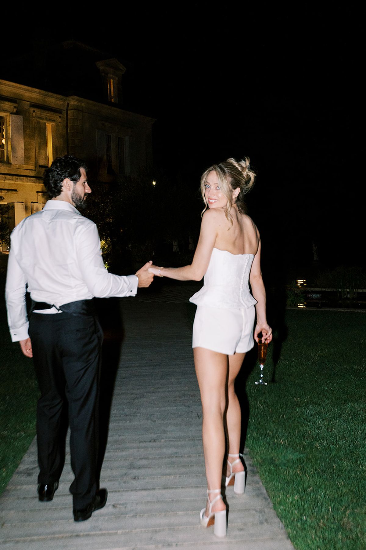 At night, the groom in shirt and dark trousers holds hands with the bride in a short white dress as they walk away.