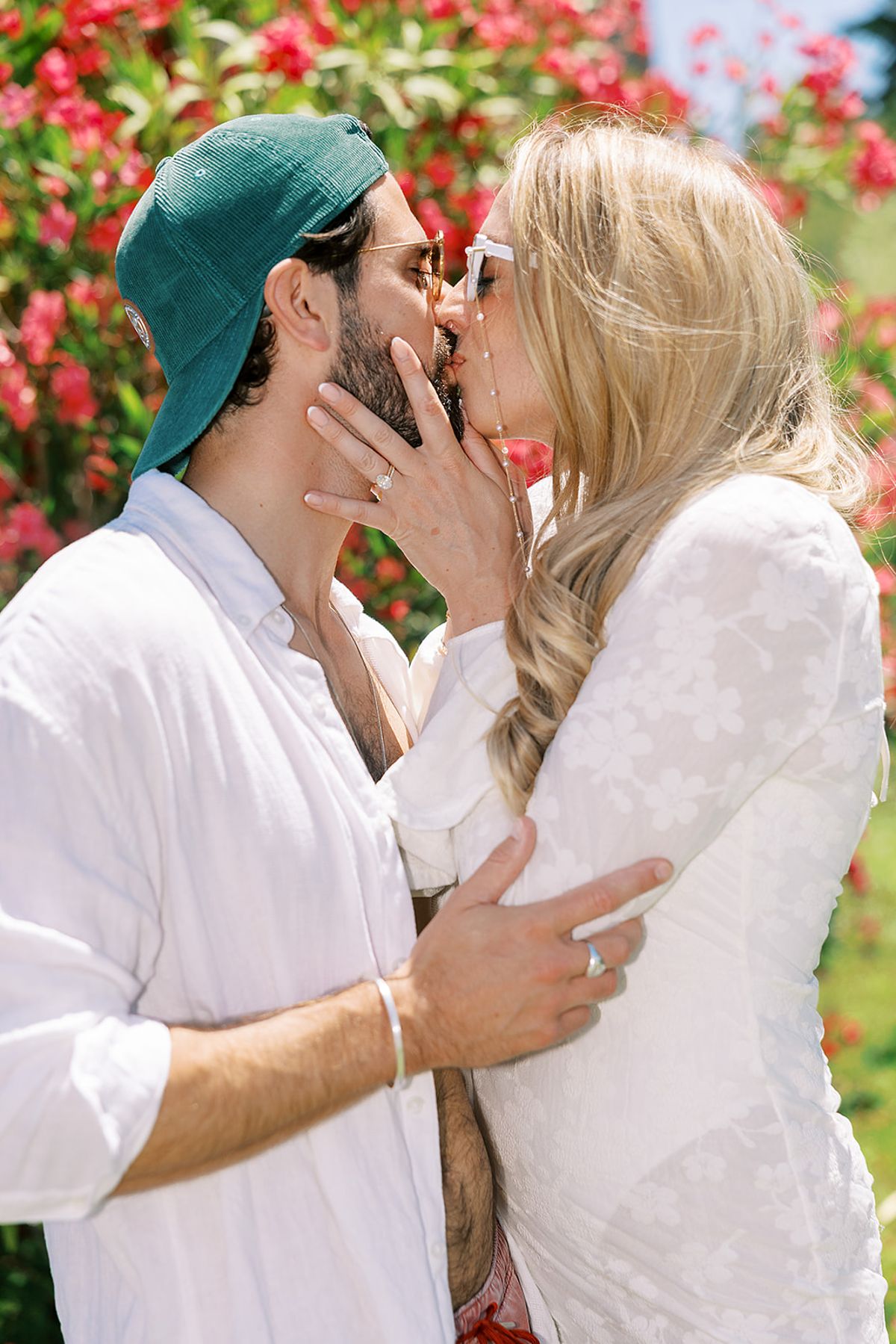The couple kiss in close-up, the groom wearing a green cap and white shirt, the bride in a white lace dress, with pink blooms behind.
