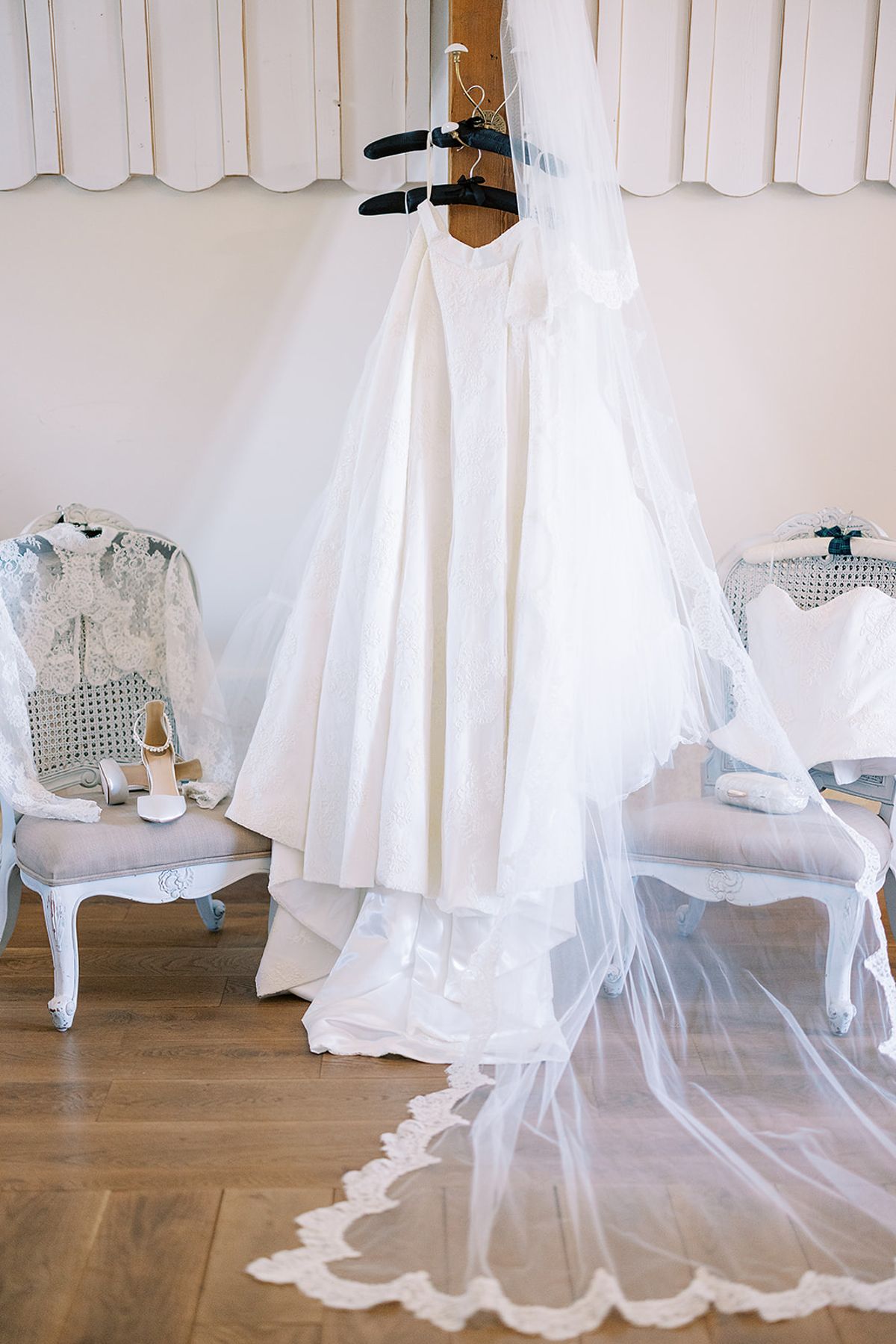 A white wedding dress hangs on a door beside a long lace-trimmed veil that trails across a wooden floor.