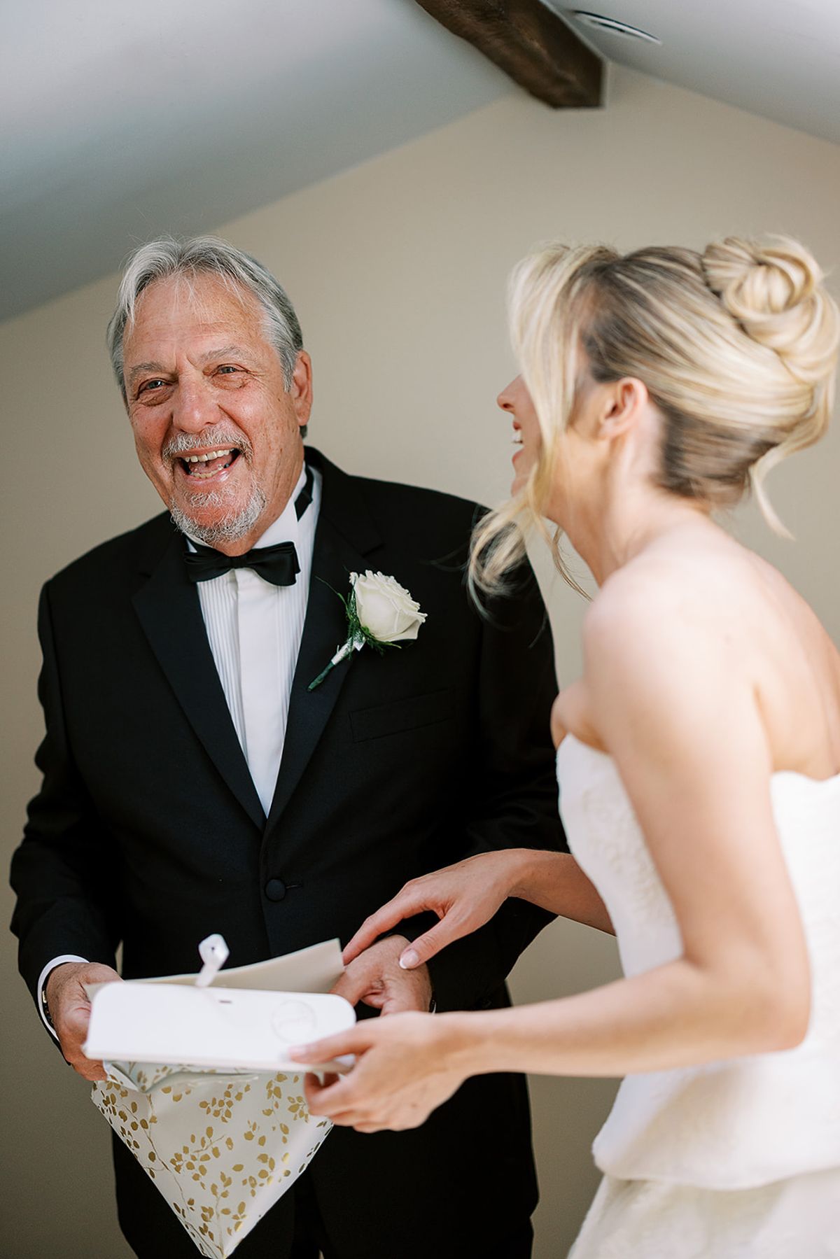An older man in a dark suit laughs with the bride as they hold a small folded parcel together.