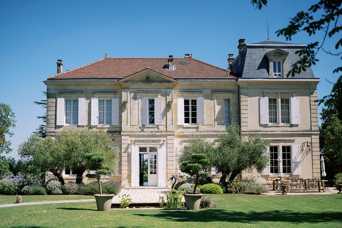 A symmetrical stone chateau with a slate and tiled roof stands behind clipped trees and a sunlit lawn.