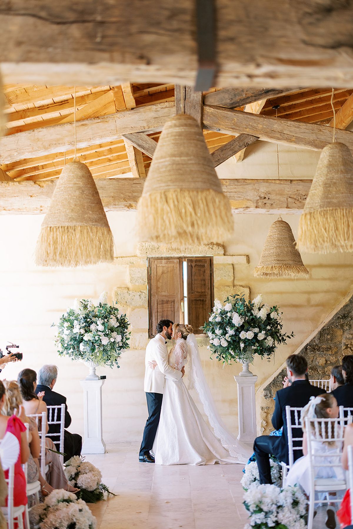 The couple kiss before white floral arrangements as seated guests look on, under rustic wooden beams and straw lampshades.