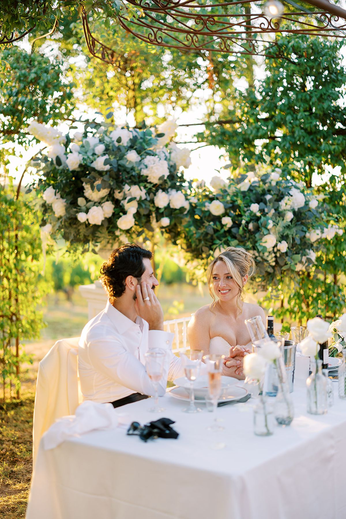 The couple sit at a white-clothed table beneath a hanging canopy of white flowers and foliage, smiling in dappled light.