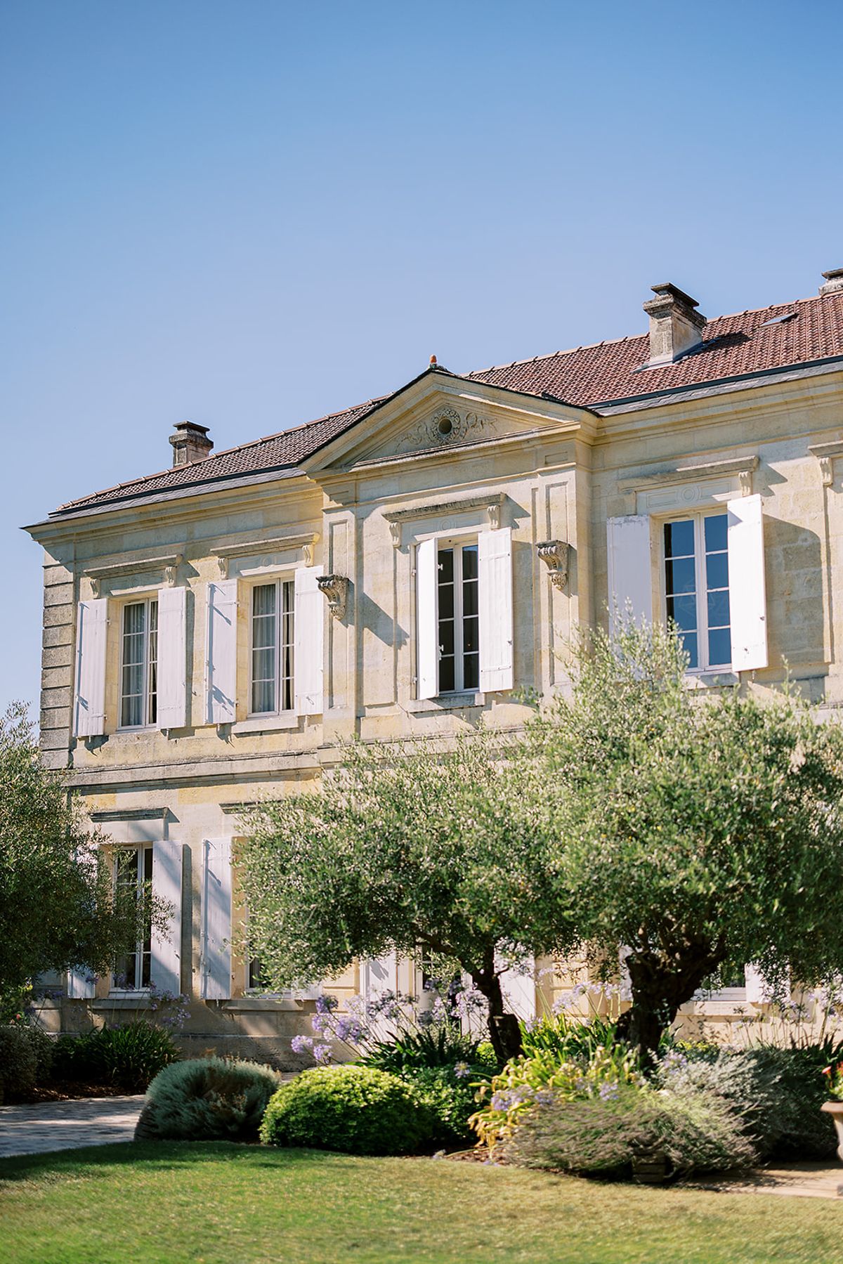 A pale stone chateau with shuttered windows and a red-tiled roof stands behind an olive tree and neat lawn.