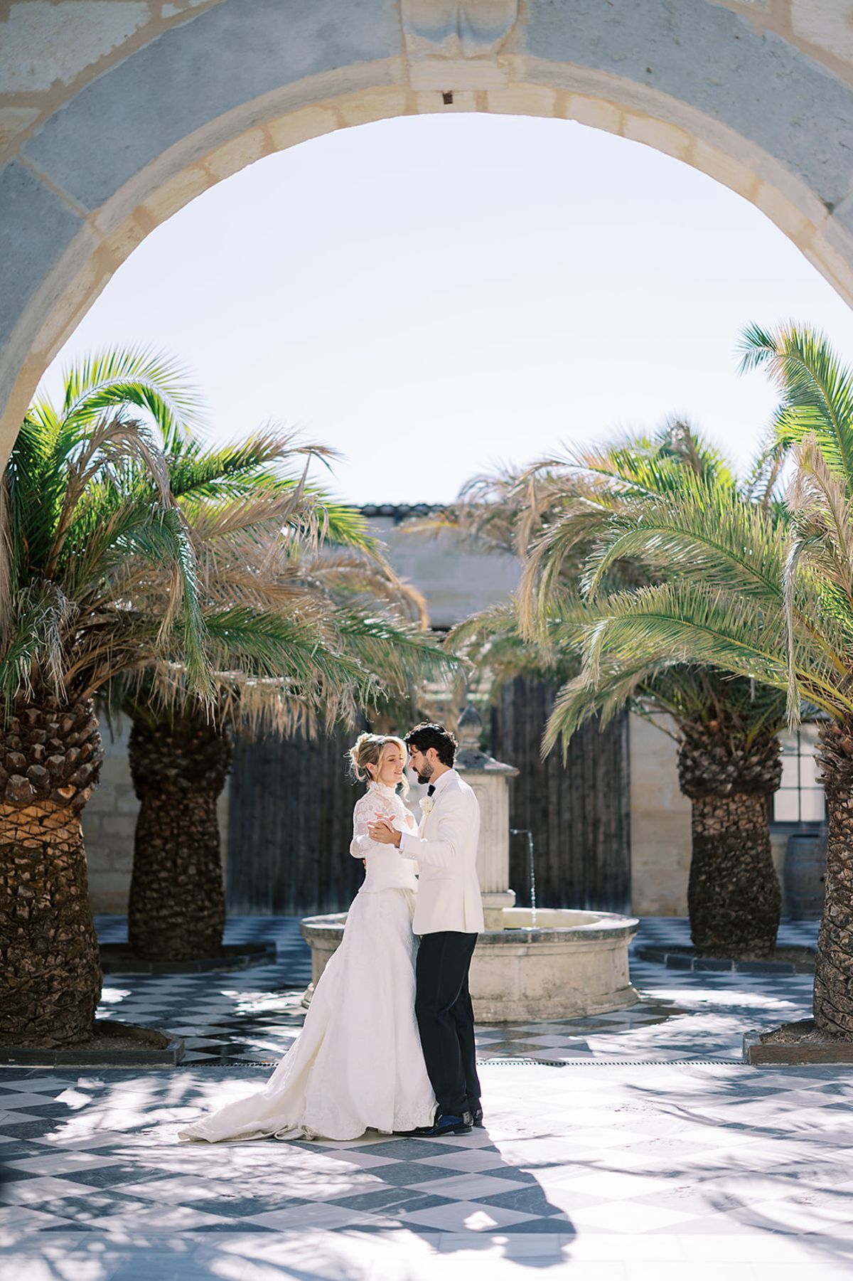 The couple embrace beside a stone fountain, framed by a stone archway and tall palm trees under a clear sky.