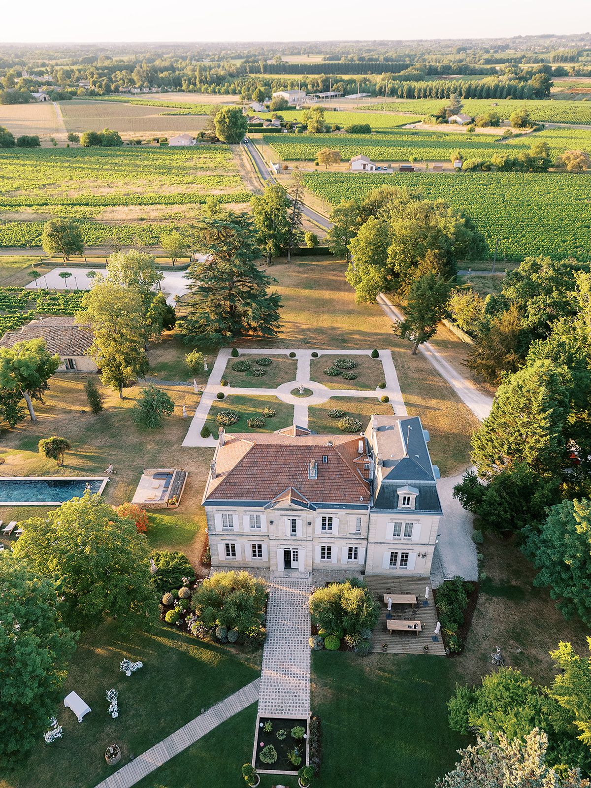 An aerial view shows a chateau with a formal garden, a swimming pool, tall trees and vineyards stretching beyond.