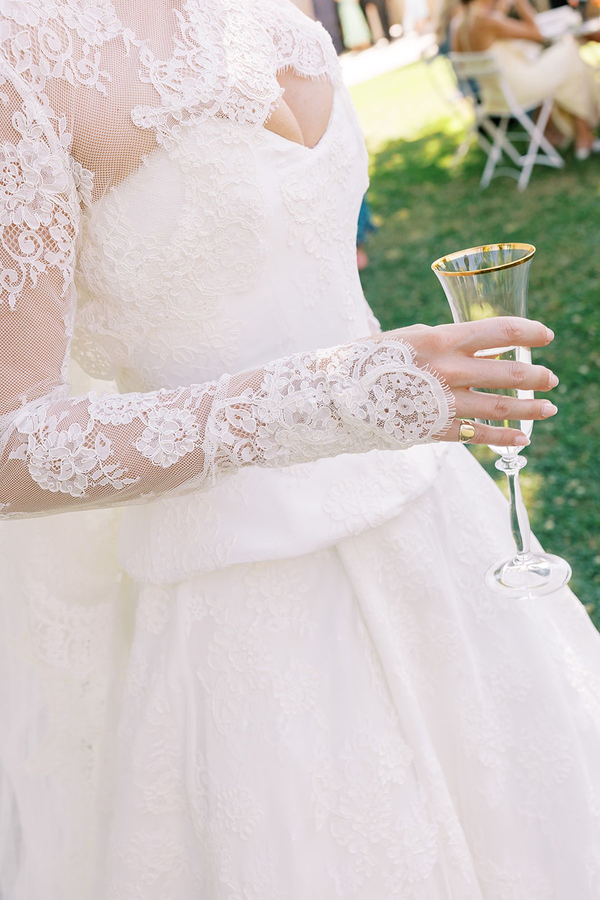 Close view of a lace long-sleeved bodice and full skirt, with one hand holding a champagne flute and guests blurred in the background.
