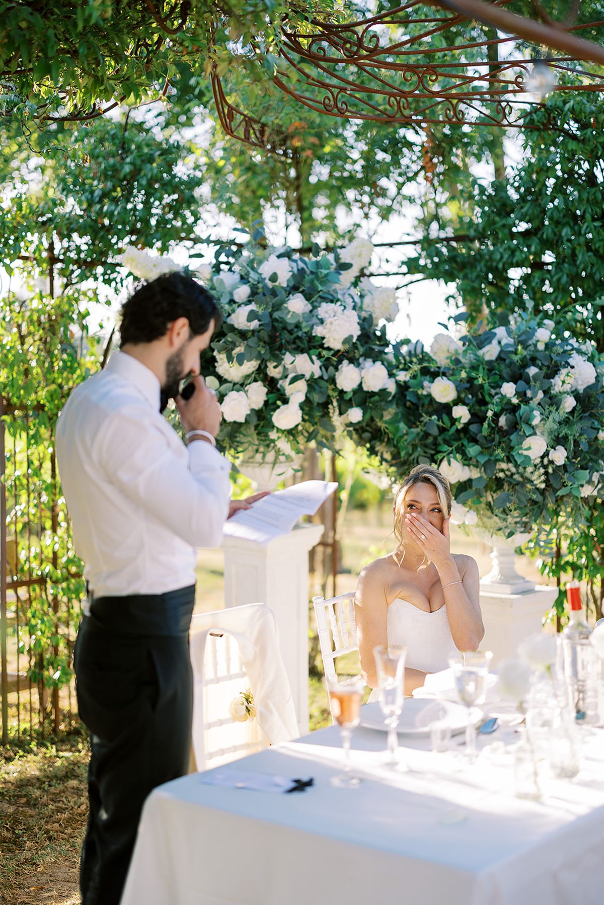 The groom reads a speech with his hand at his face while the bride laughs at a table under a flower canopy.