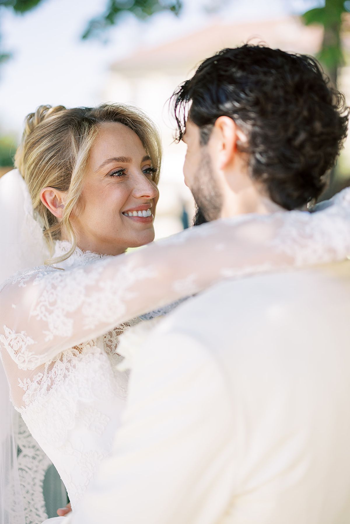 The smiling bride, wearing a lace-sleeved dress, looks at the groom as they embrace in bright natural light.