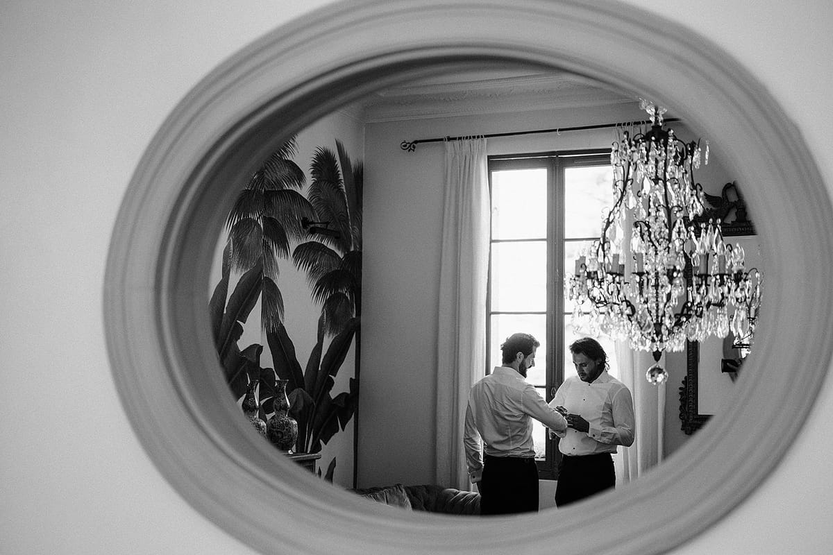 In black and white, an oval mirror reflects two men standing close together by a tall window and a crystal chandelier.