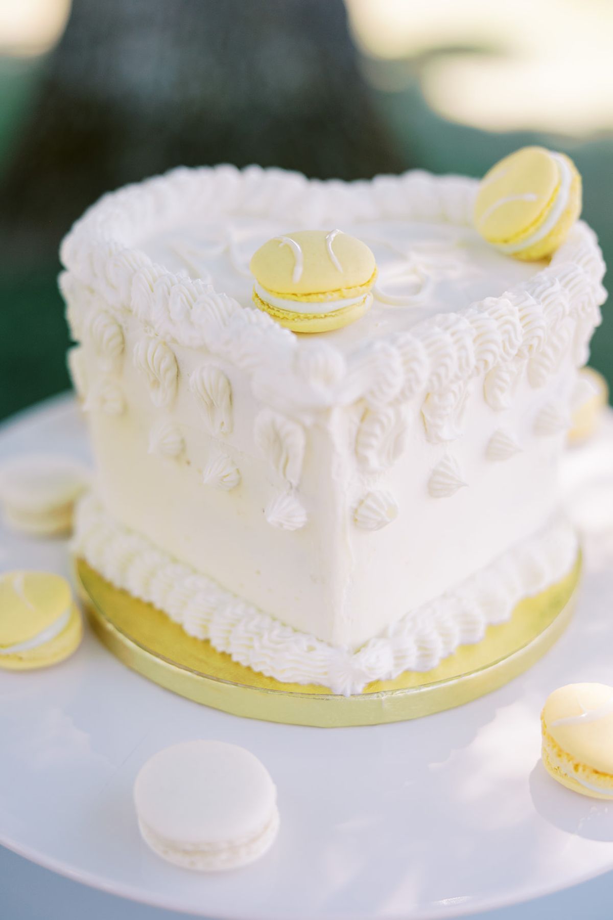 A heart-shaped white cake with piped frosting is topped with pale yellow macarons, more macarons around its base.