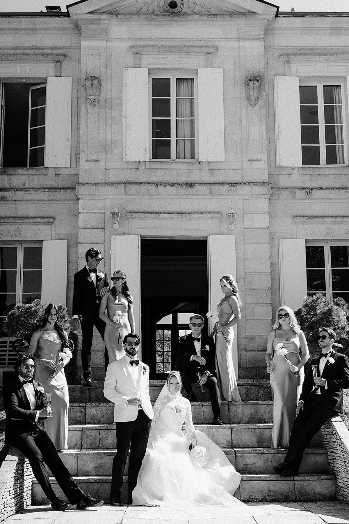 The couple sit on stone steps at the chateau doorway, surrounded by their wedding party standing and sitting around them.